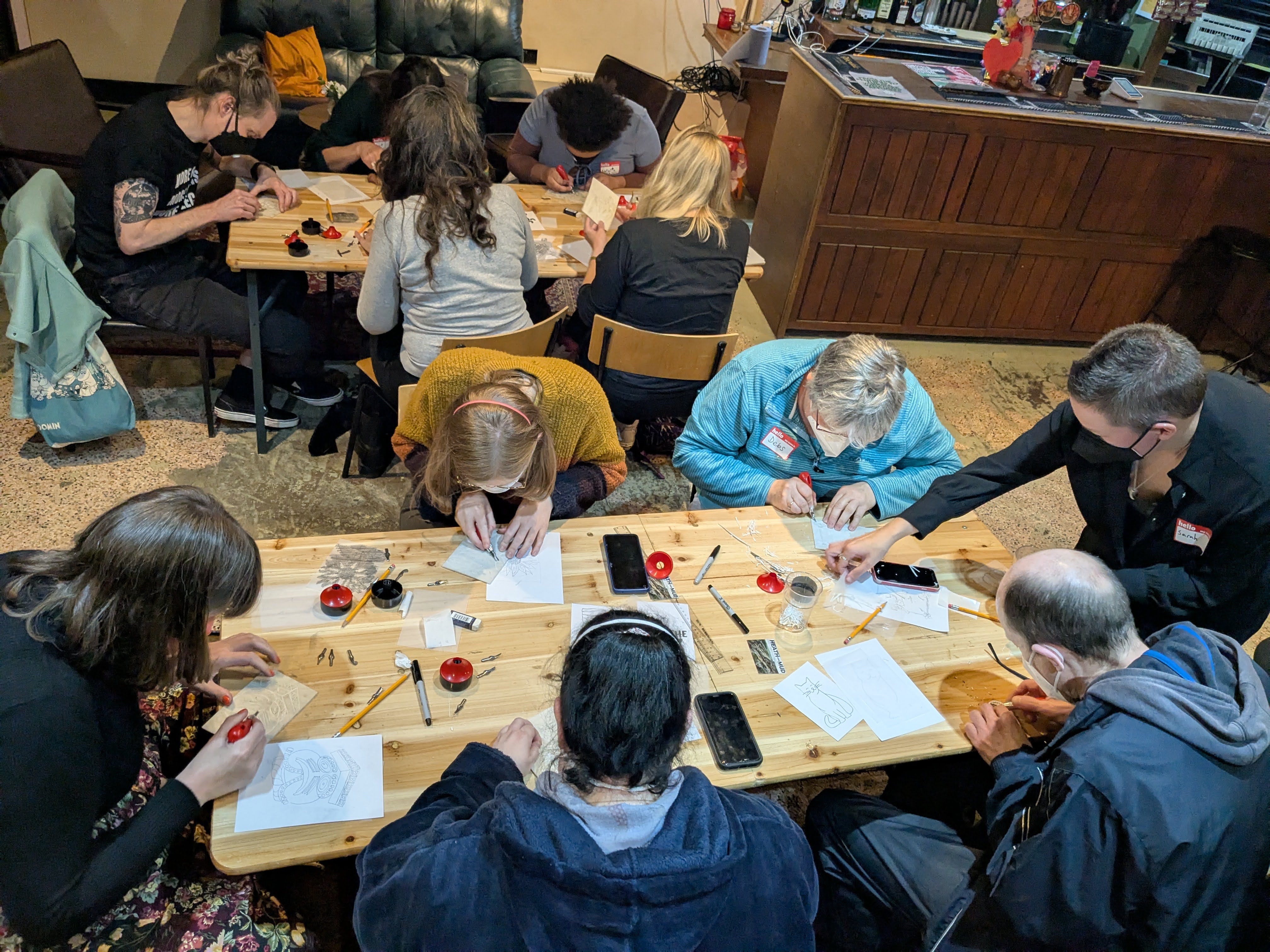 Two tables of masked people are being instructed in lino cutting techniques
