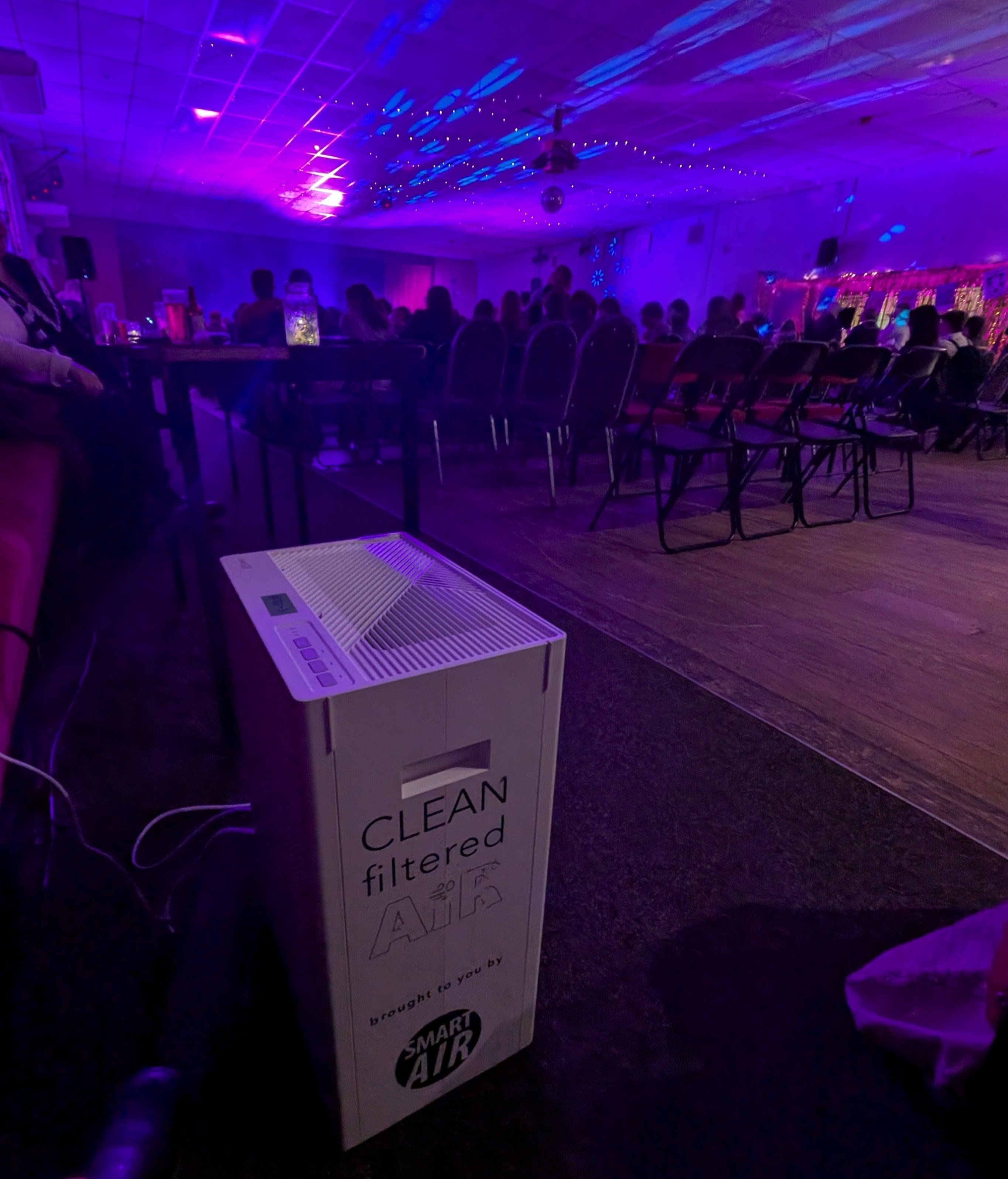 An air purifier at the back of an event space. There are rows of chairs in front of the stage and the lighting makes everything look purple and blue