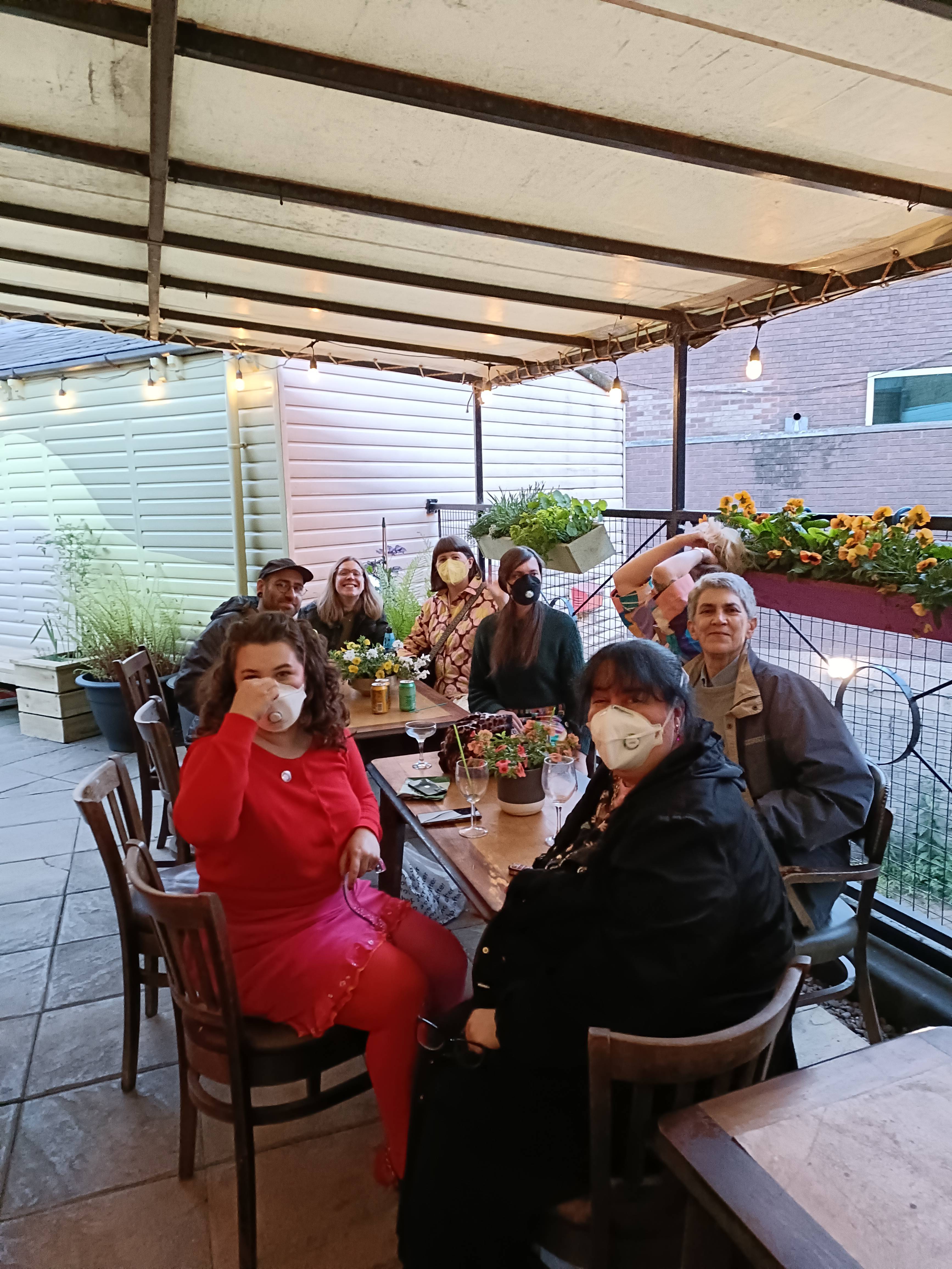 A group of people outside sitting around a table, most people are wearing masks and there are flowers in the background in nice pots